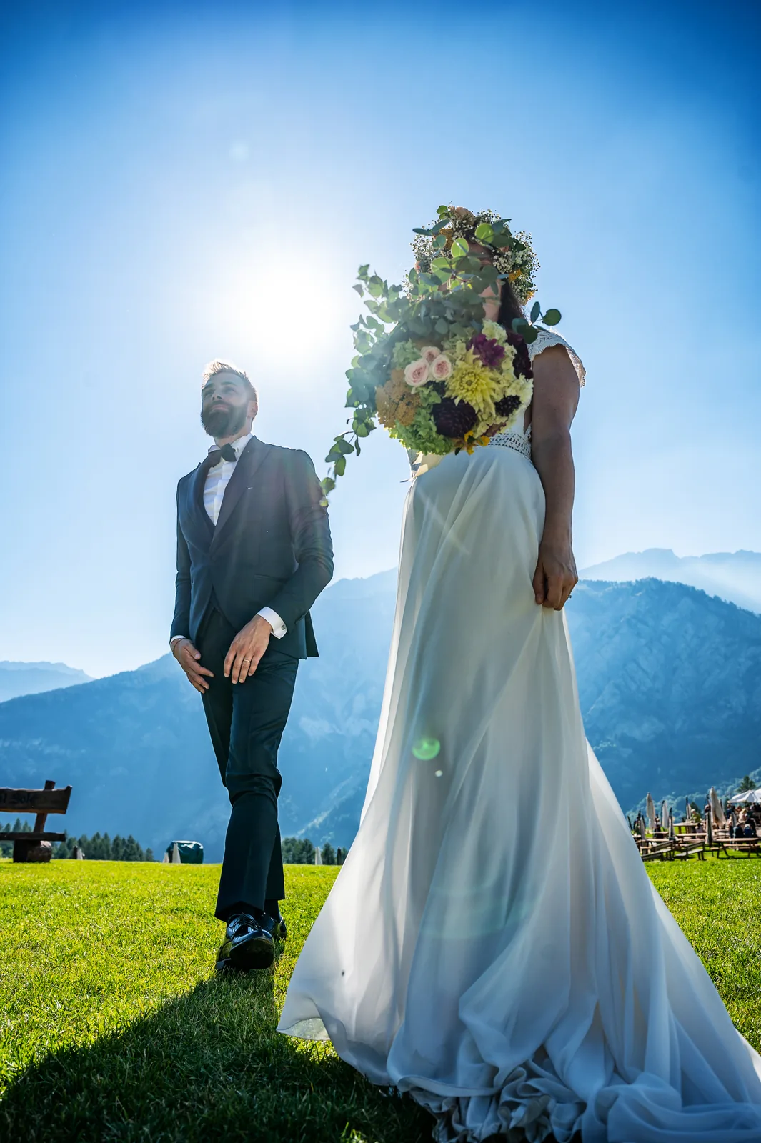 The bride raising her bouquet toward the sun beside the groom, Alpine valley behind