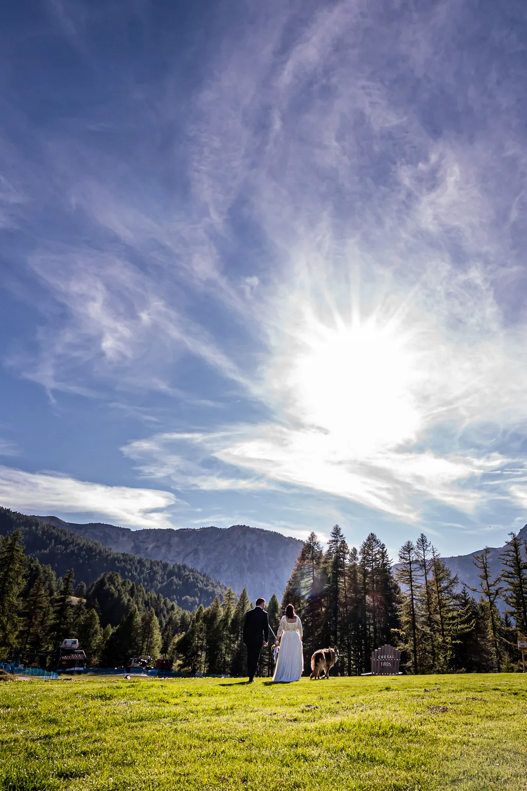 A vast sky with the sun above a tiny couple on an Alpine meadow