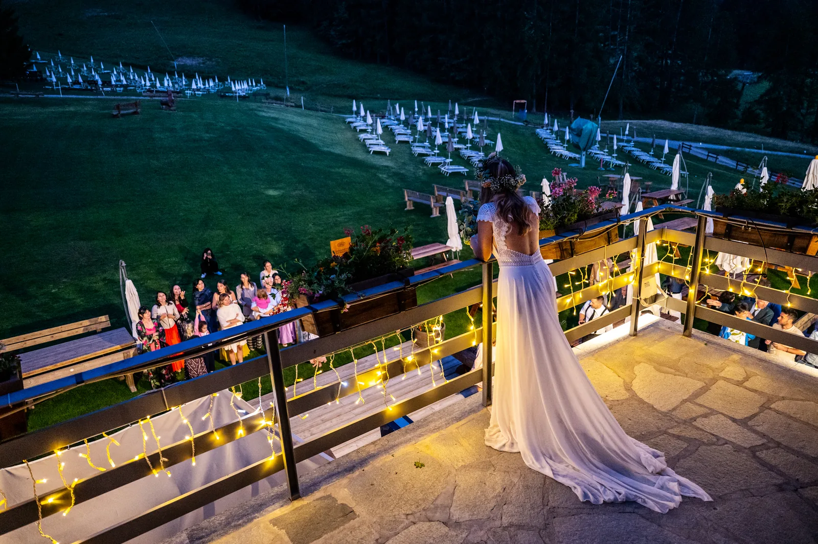 The bride on a mountain terrace at dusk, looking down at her guests among string lights, Bardonecchia
