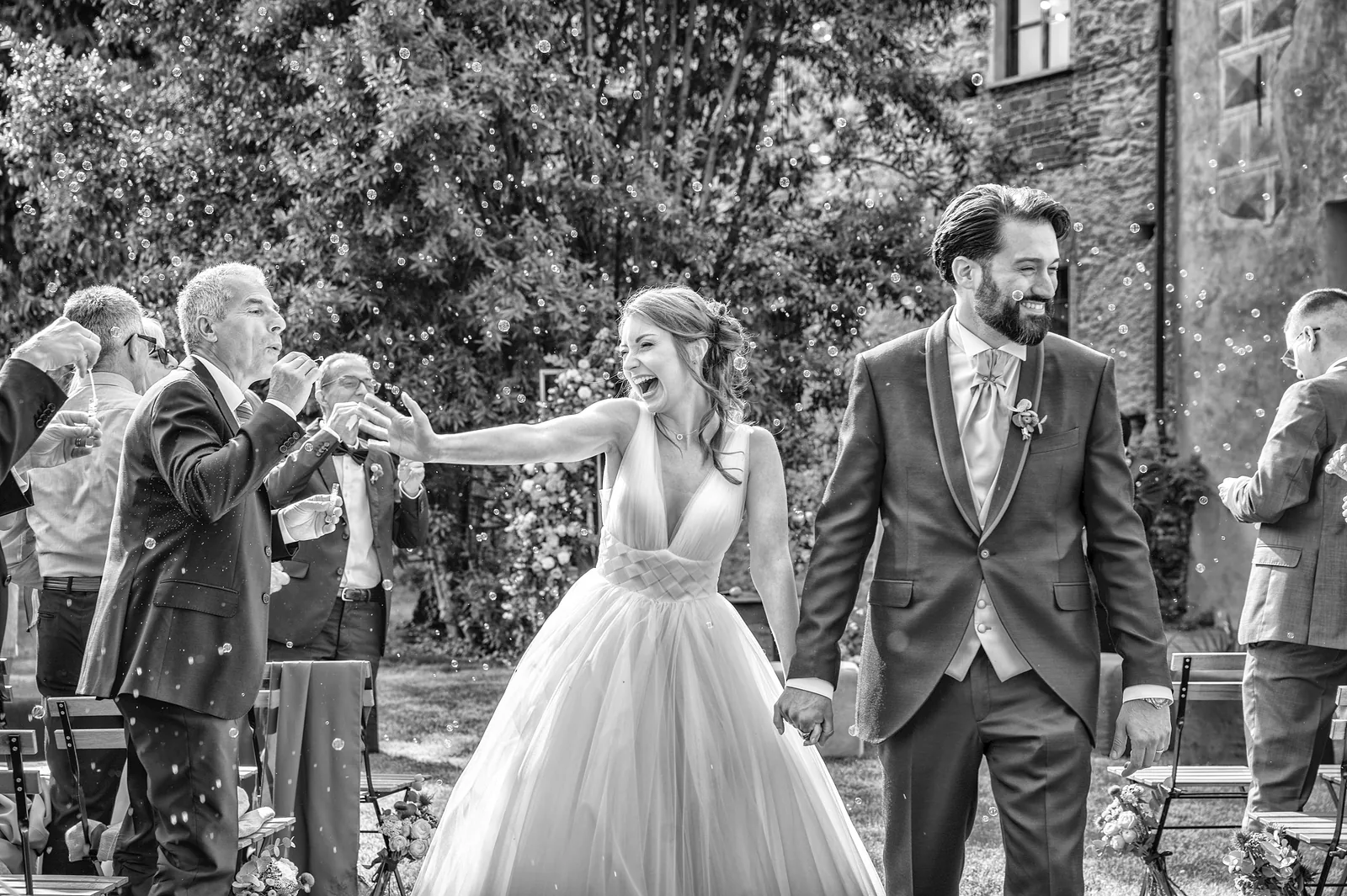 The bride laughing and reaching toward a guest as the couple walk out of the ceremony, Castello della Costa, black and white