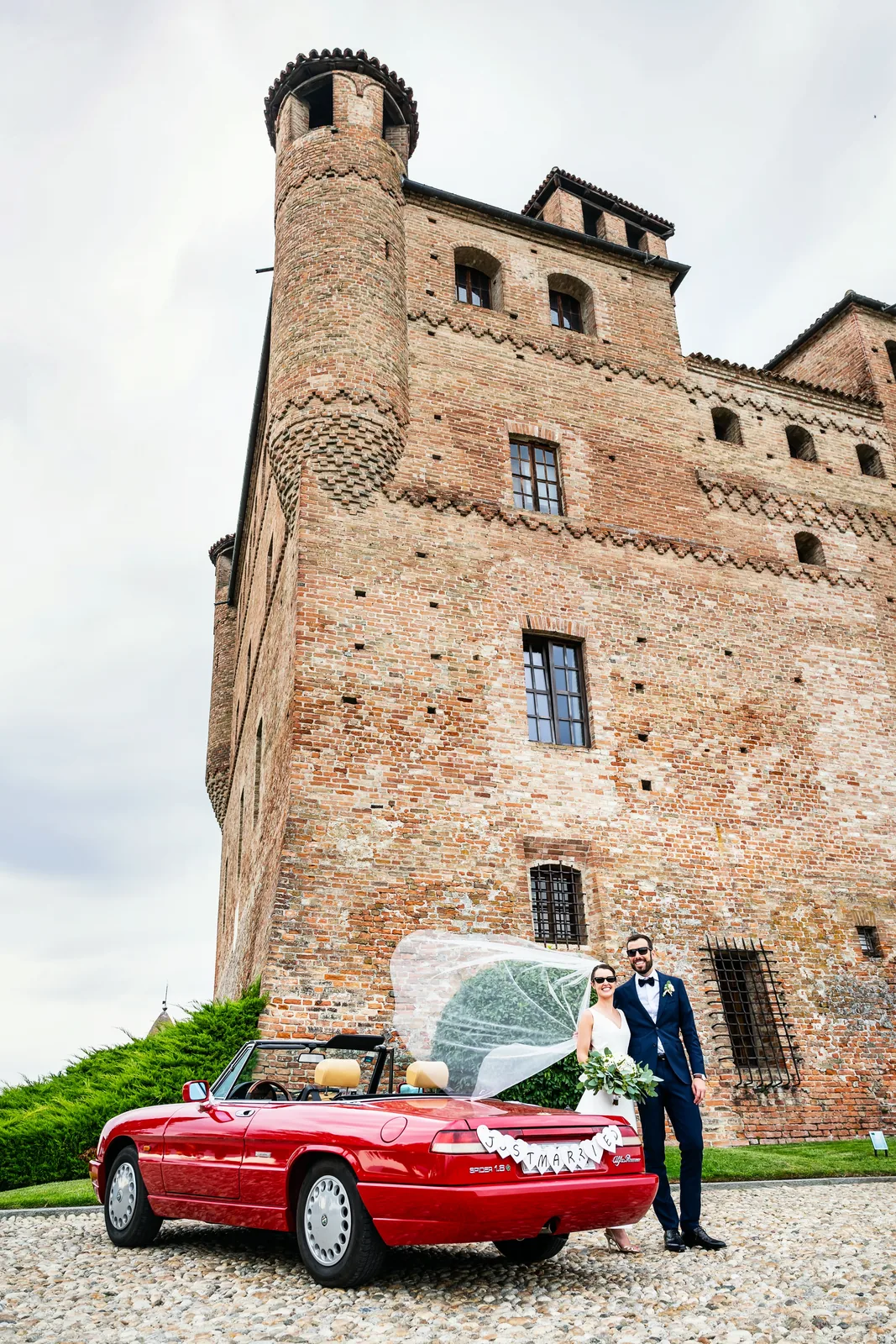 The couple beside a red vintage Alfa Romeo under the tower of Castello di Grinzane Cavour