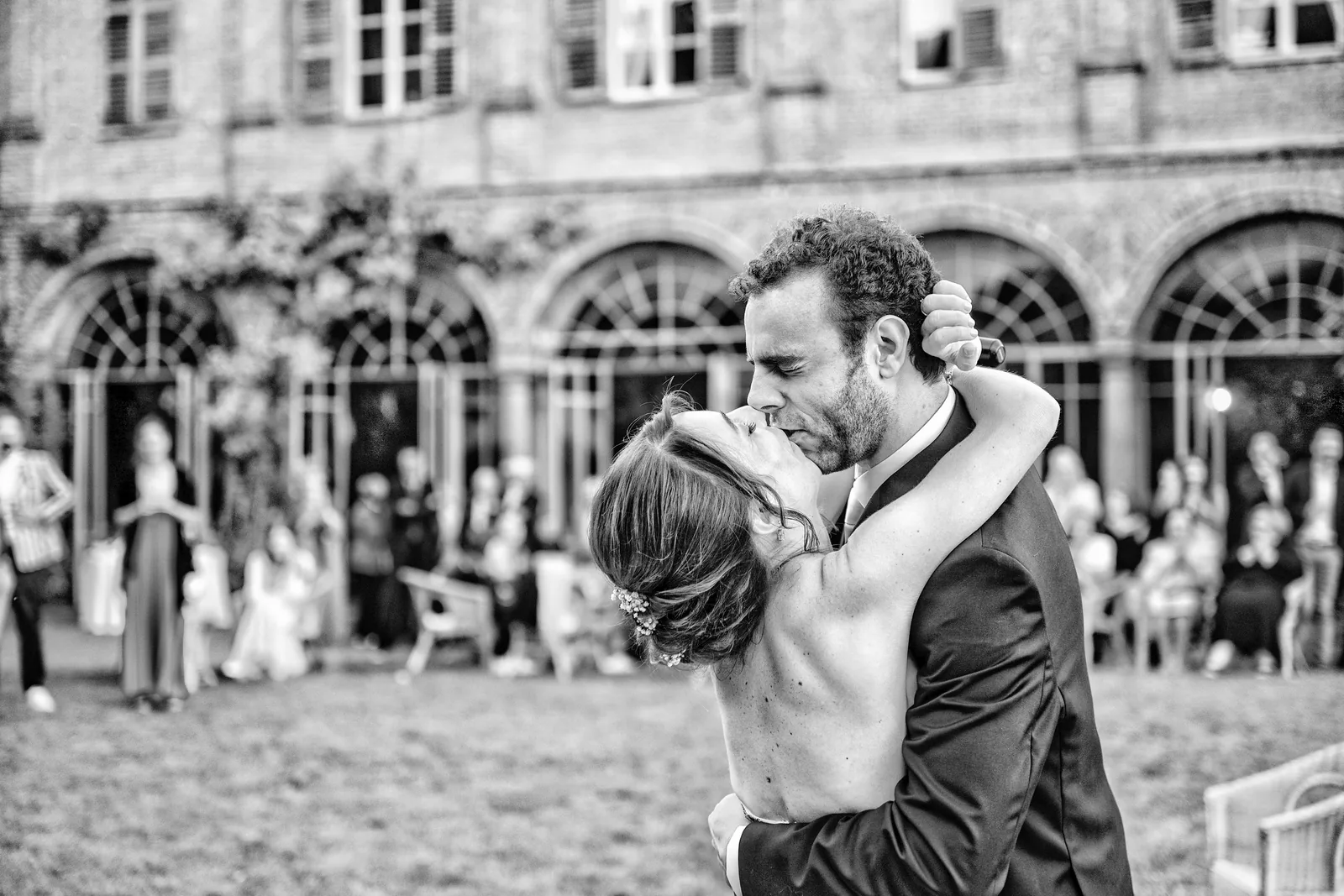 The couple kissing in an arcaded courtyard as guests watch, Palazzo La Marmora, black and white