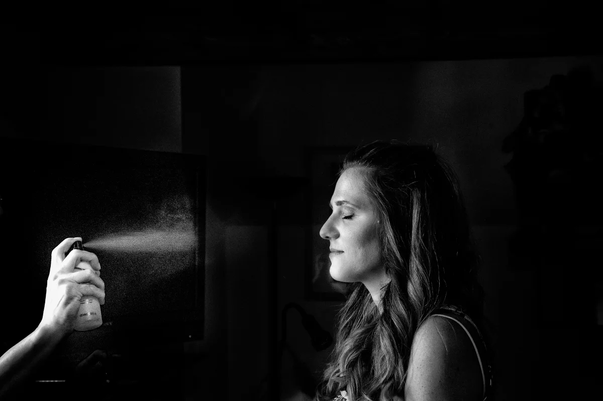 Sposa di profilo nella stanza dei preparativi, mano che spruzza profumo controluce dalla finestra, fotografia in bianco e nero di Elisabetta Rosso